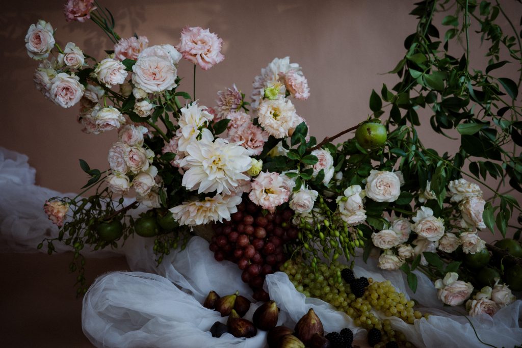 Urn arrangement filled with flowers including cafe au lait dahlias, pink garden roses, pink and peach lisianthus and lime branches, with red and green grapes and figs on white organza fabric
