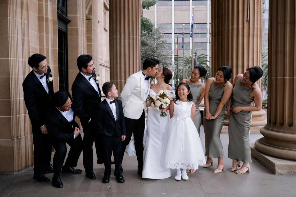 A wedding couple kiss, standing in the centre of their wedding party, to the right is the bridesmaids, three, dress in long silk olive dresses, in front of the bride stands a young flower girl. The groom stands to the left of the bride and three grooms men and a page boy are wearing black and white suits. the wedding party have turned heads to watch the couple kiss