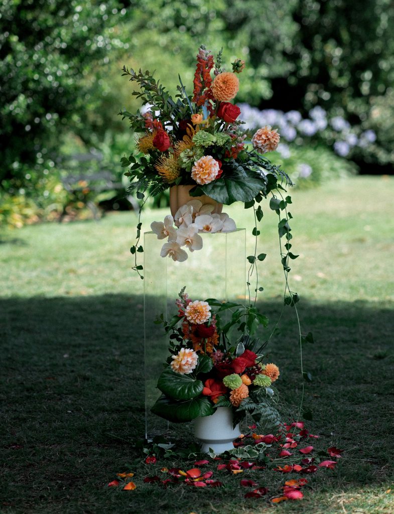 Wedding ceremony installation on clear acrylic plinths in red, orange and yellow colours, including pincushion, orchids, dahlians snapdragons, zinnia, ivy and fern foliage with rose petals on the ground