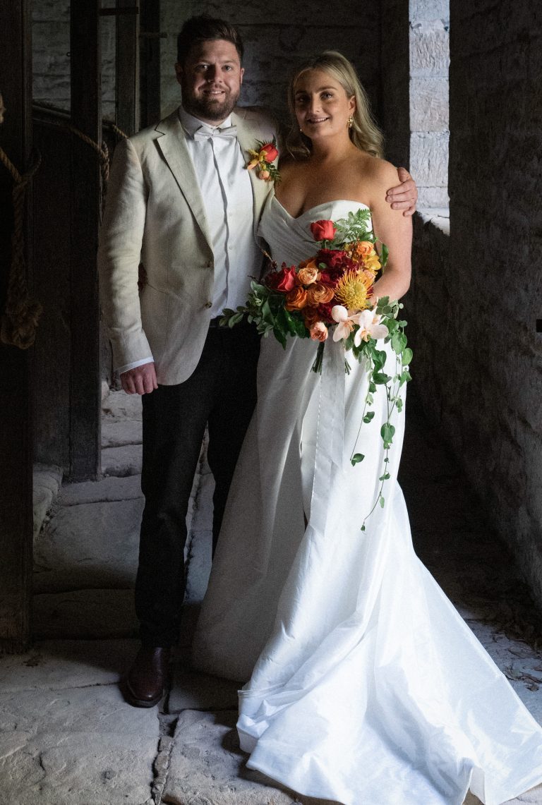 Bride and groom standing in stable with their bouquet and buttonhole in yellow orange and red colours, containing roses, native pincushions, copper and rust ranunculus, eucalyptus foliage and peach orchids