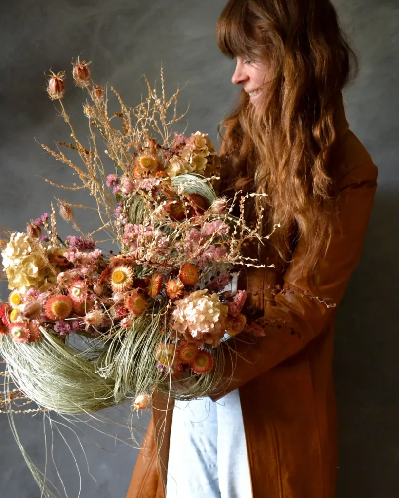A woman with long red hair stands at the right side of the frame, facing the left. She wears a long terracotta coloured jacket and white pants, she is smiling and looking downward at a dried floral arrangement she holds. The flowers are dried grasses, with earthy toned dried straw flowers, in yellows, and oranges. There is also dried hydrangea and thistle.