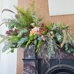 A large floral design, lots of Australian native foliage, sits on the corner of a fire place mantle