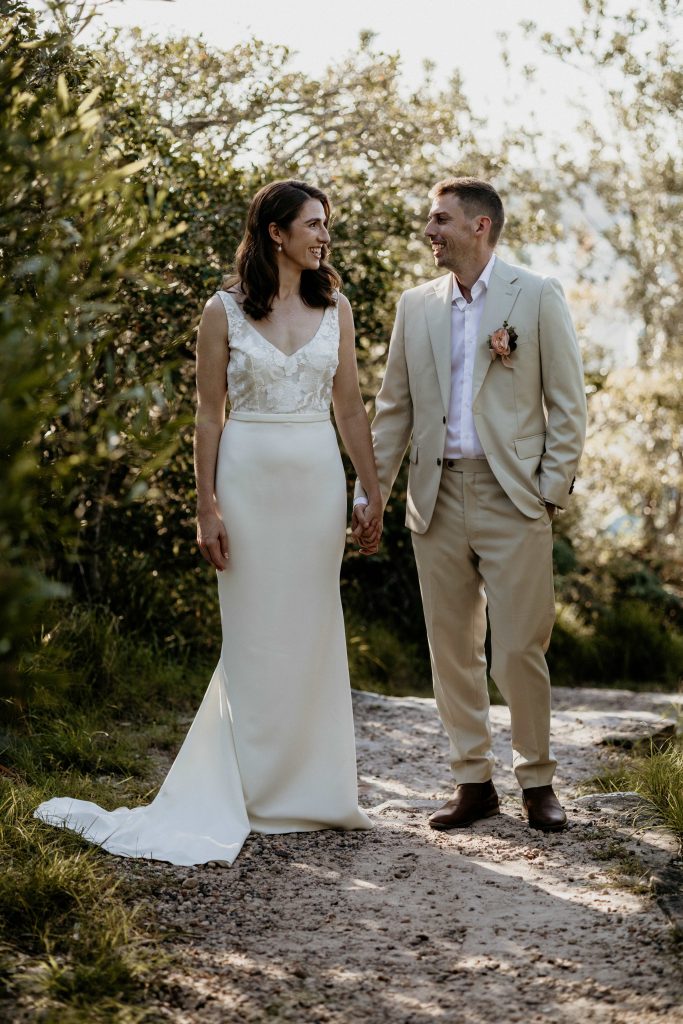 A bride and Groom stand in full length image, side by side, looking at each other smiling. The bride has deep red hair and a long ivory gown. The groom wears an oatmeal coloured suit and white shirt. They stand in an outdoor setting, on a sandy path