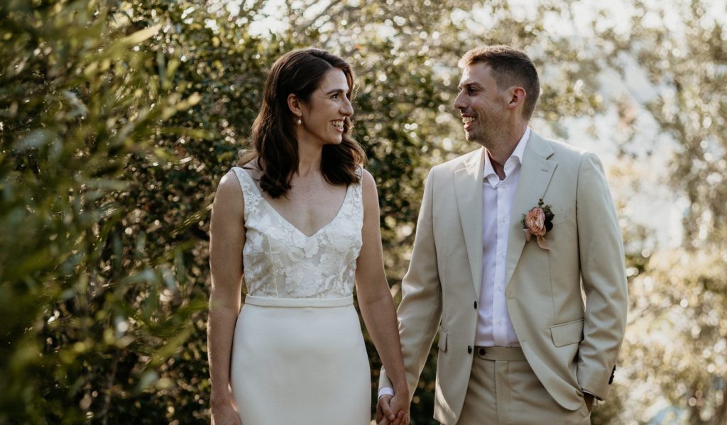 A bride and Groom stand in full length image, side by side, looking at each other smiling. The bride has deep red hair and a long ivory gown. The groom wears an oatmeal coloured suit and white shirt. They stand in an outdoor setting, on a sandy path