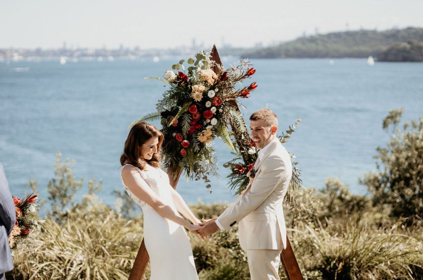 A wedding couple stand in front of a floral arch holding hands during their ceremony. There is ocean in the background.