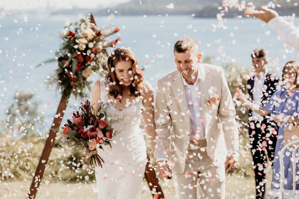 A newly married couple walk down the aisle, rose petal confetti fills the air as they walk past their guests.