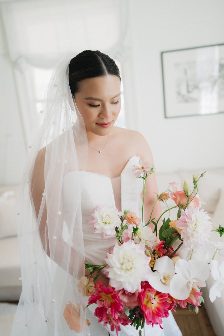 A bride all in white, looks down at her bouquet. Her bouquet is white, pink and a hint or orange.