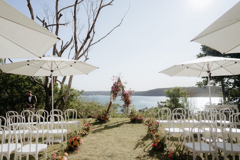 An A frame arbour covered with pinks, oranges and white flowers sits in the background of a wedding aisle. The ocean can be seen on the horizon, the sun is low