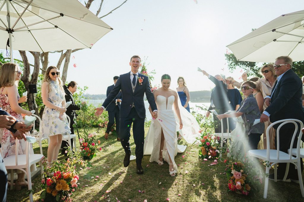 A newly married couple walk hand in hand down the aisle, guests on either side clapping and cheering