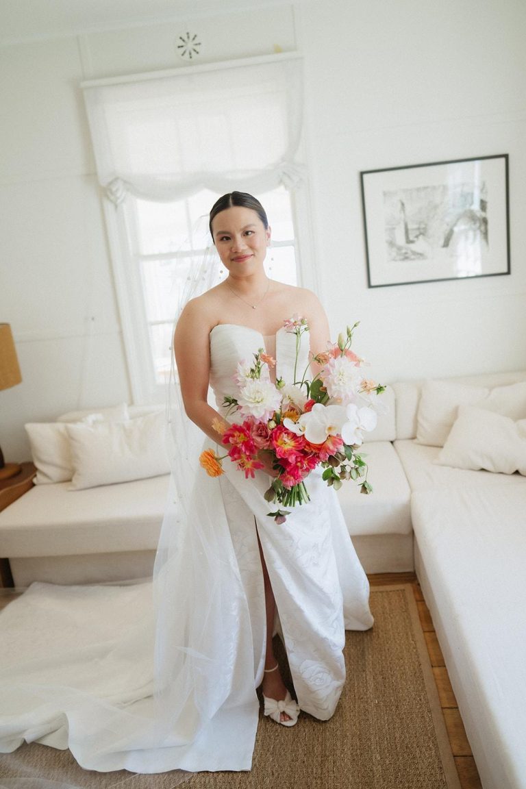 Full length image of bride in floor length white, holding her bouquet in front of her. Her bouquet is white, pink and orange