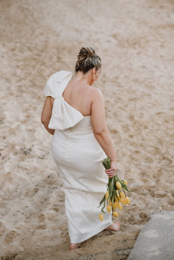 A bride in a white dress, with a large bow on her back left shoulder, walks down the sand bank with yellow tulip bouquet in hand
